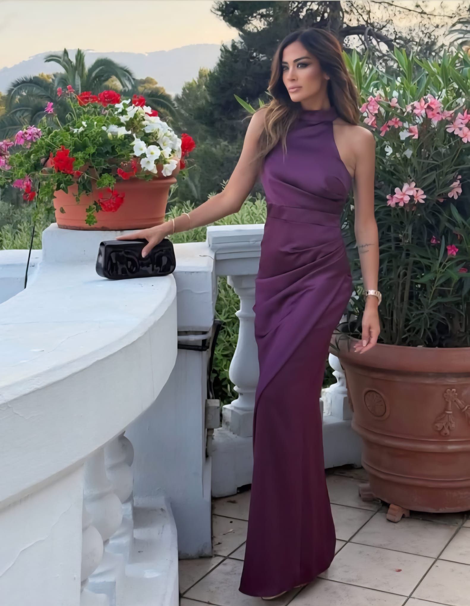Paarse satijnen halterjurk met een zwarte clutch op een terras met oleanders.