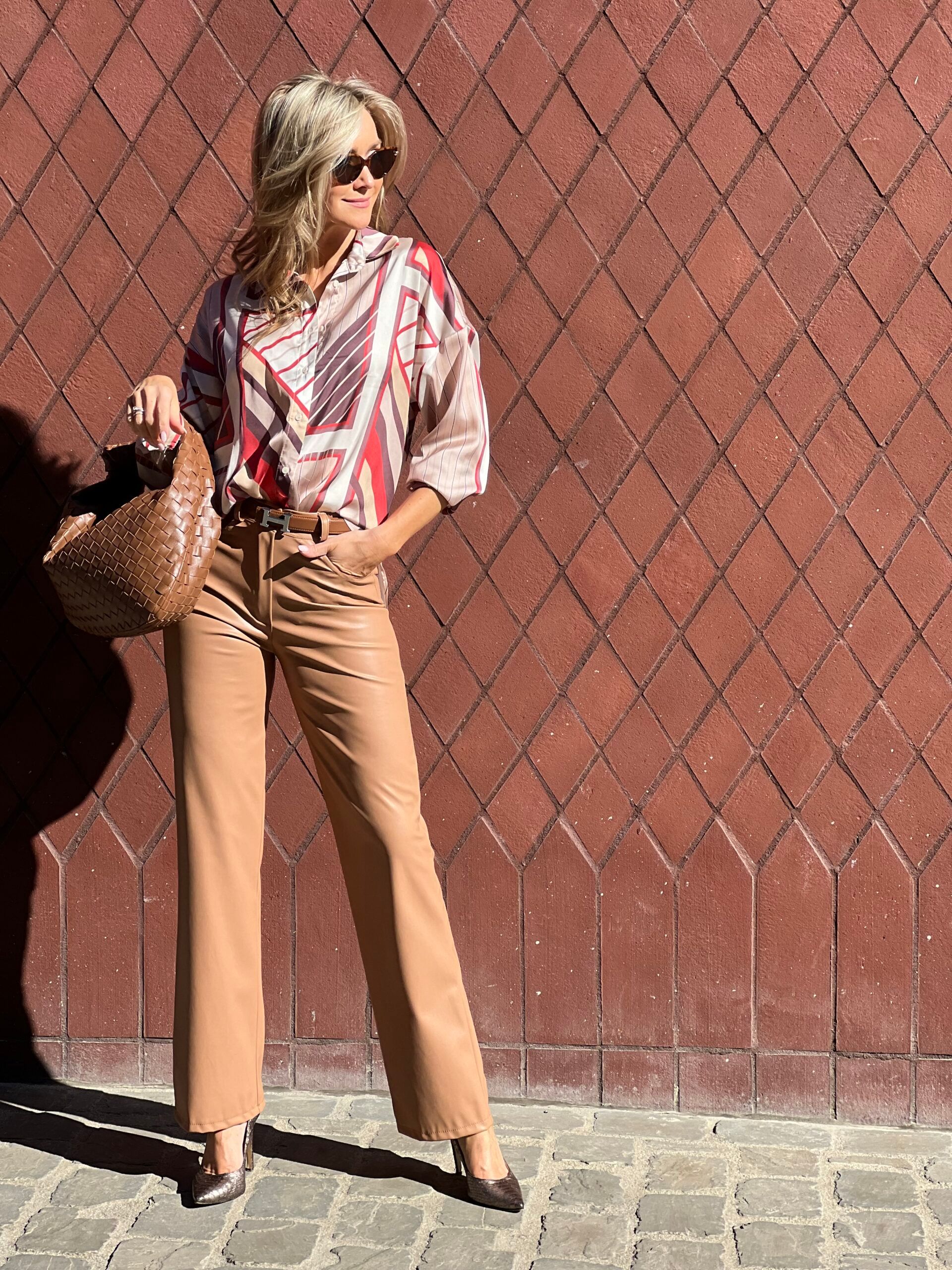 Blouse met print, camelkleurige broek, pumps en gevlochten tas op een kasseiweg.