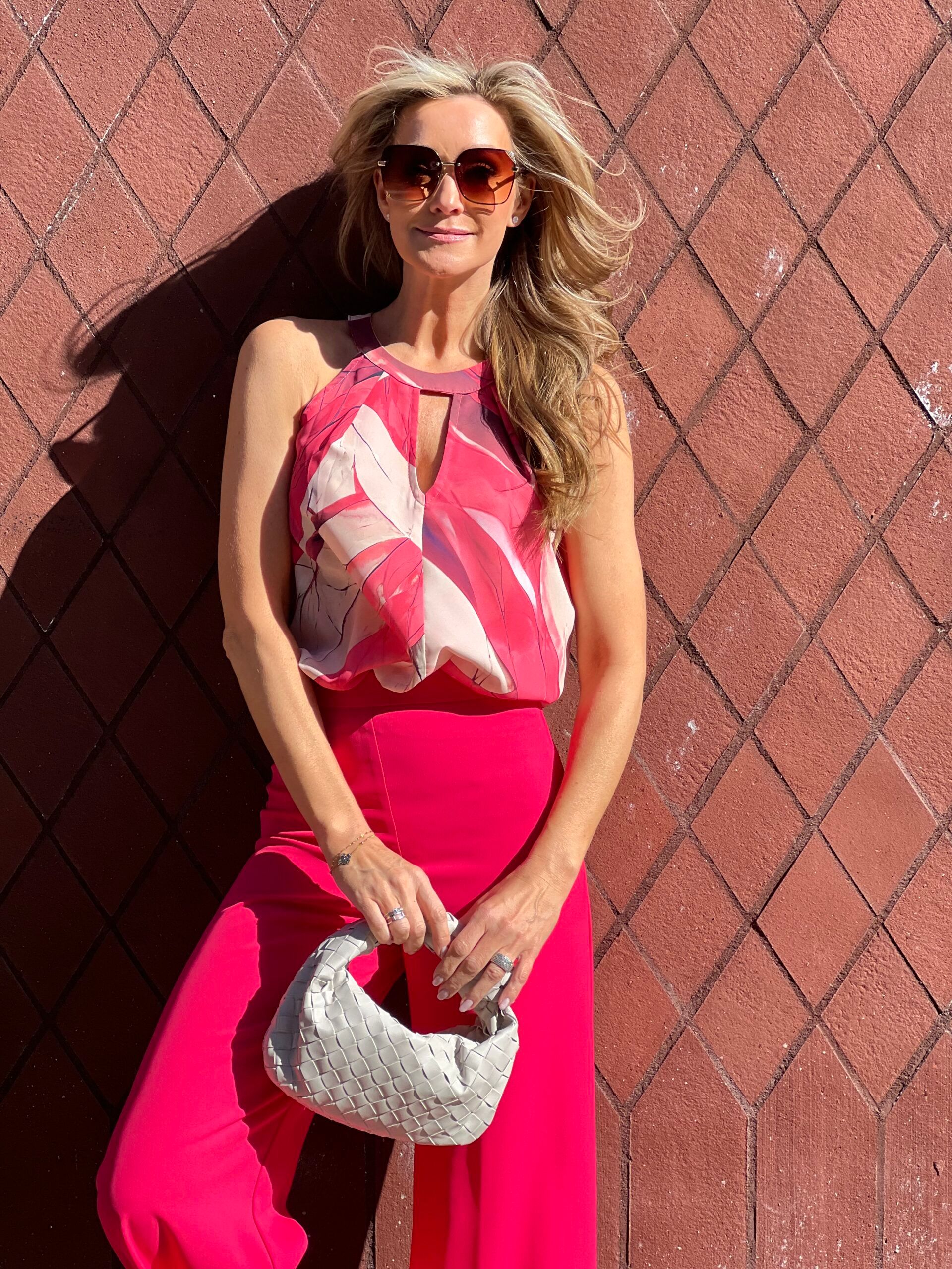 Fuchsia top met print en fuchsia broek met een witte gevlochten tas tegen een bakstenen muur.