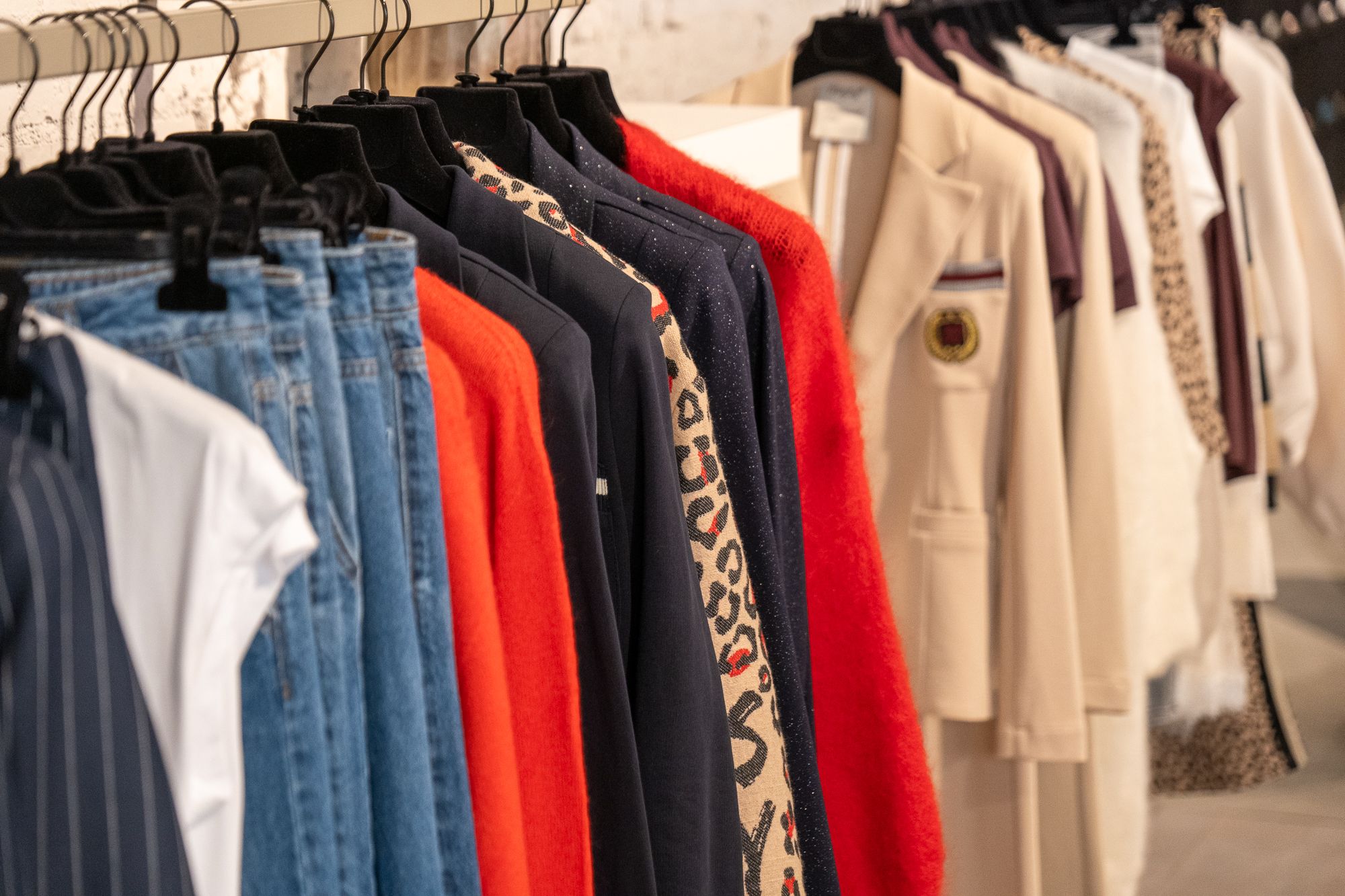 Kledingrek met denim, een rode trui, wit-blauwe blouses en een roomkleurige blazer.