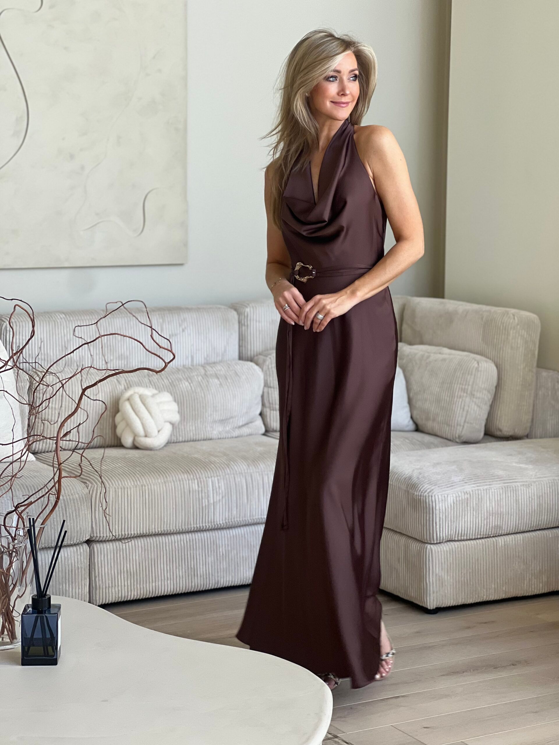 Vrouw in een chocoladebruine satijnen maxi-jurk met ceintuur, staand in een lichte woonkamer.