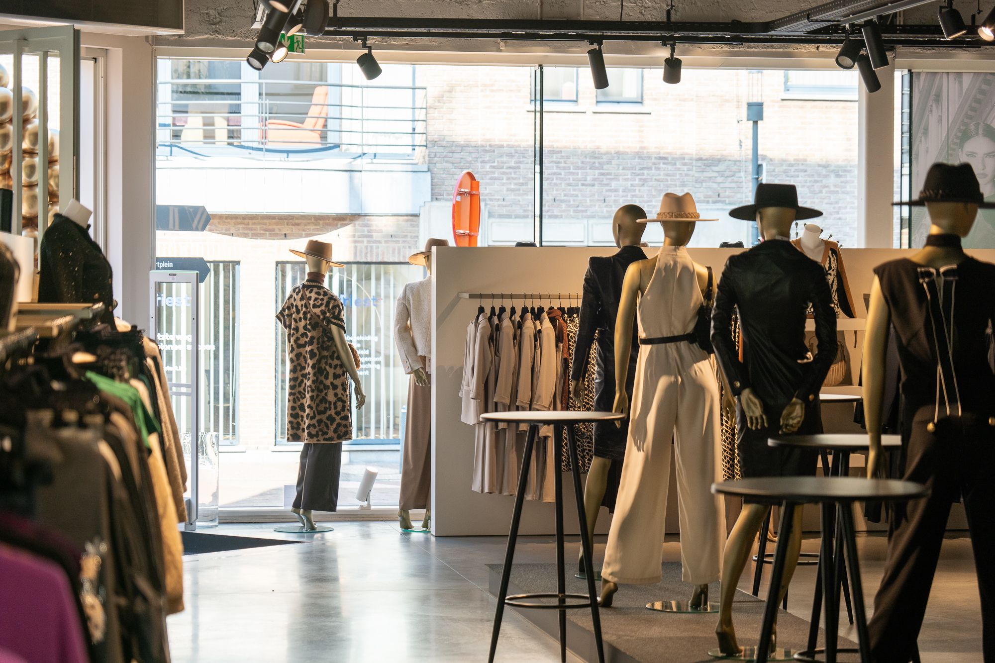 Overzicht van de boetiek: kledingrekken, gouden paspoppen op een podium en klanten bij de vitrine aan de straatkant.